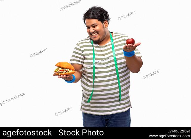 Portrait of a fat man with measuring tape around his neck greedily devouring a burger rather than apple