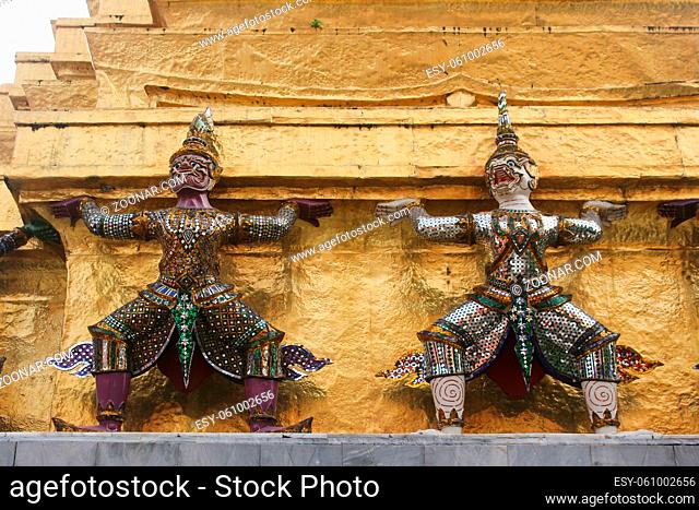 Horizontal close up photo of two demon statues, or demon guardians, holding Golden Chedi prang at Wat Phra Kaew temple. Bangkok, Thailand