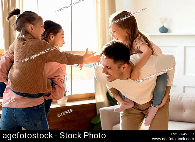 Happy family young adult parents piggyback little children daughters playing together in living room, cheerful mum dad carrying cute small kids on back having...