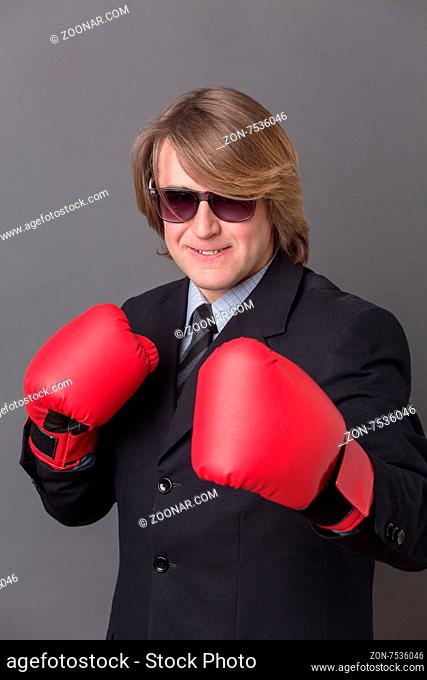 Middle-haired businessman smiling in red boxing gloves. Young man in sunglasses ready to compete with his business rivals