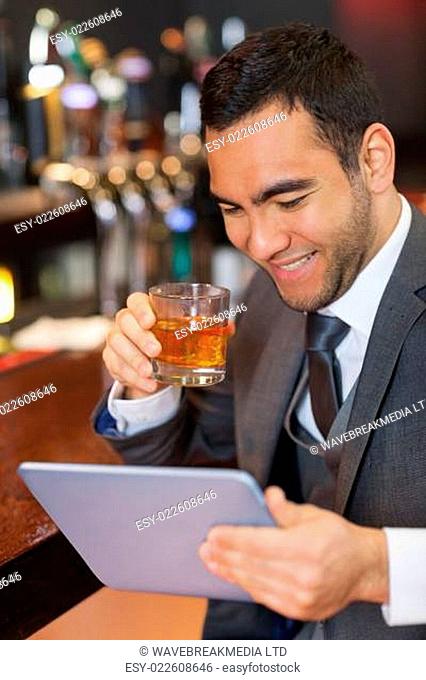 Smiling businessman working on his tablet while having a whiskey