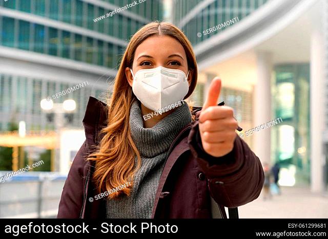 Confident woman wearing protective mask KN95 FFP2 showing thumb up and looking at camera in modern city