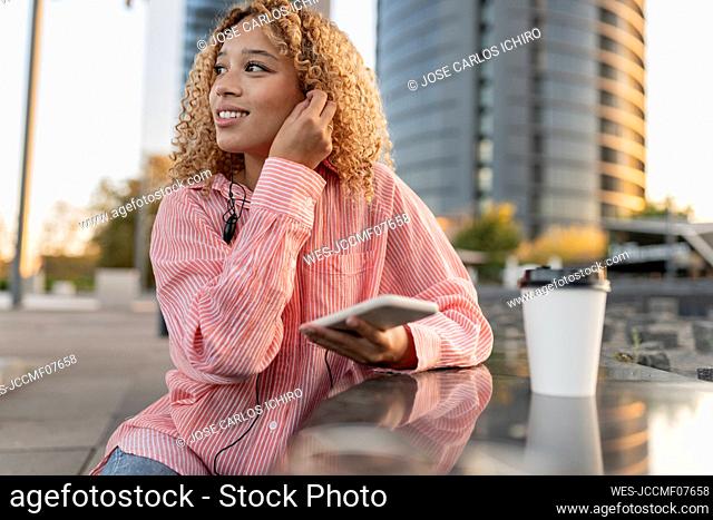 Smiling woman with smart phone wearing in-ear headphones by bench