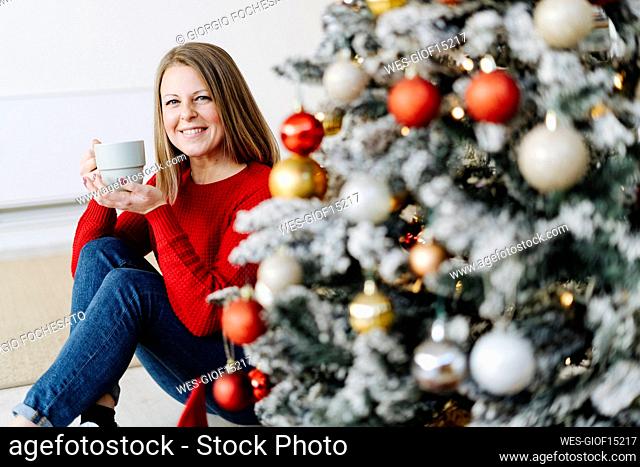 Happy woman with coffee cup sitting by Christmas tree at home