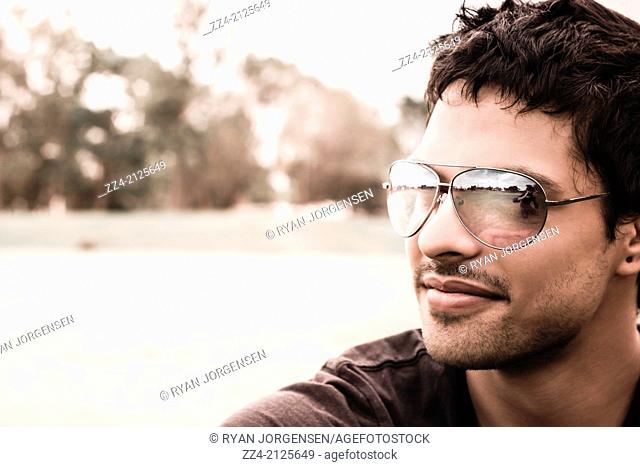 Horizontal portrait of a handsome Hispanic man in mid-twenties relaxing at an outdoor park location wearing sunglasses