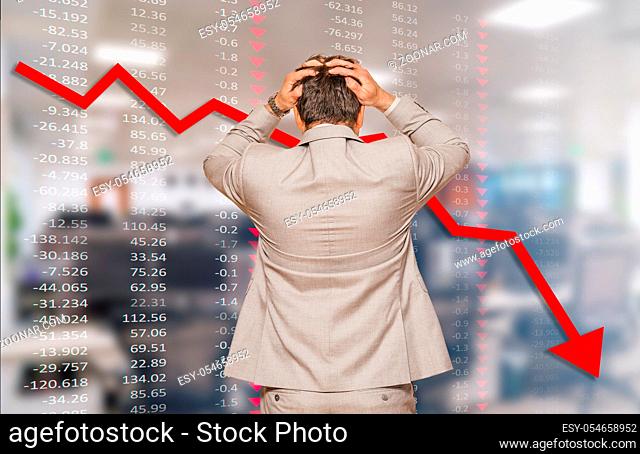 Back view of middle age businessman pulling his hair out as his business falling during recession, data chart and blurred office in background