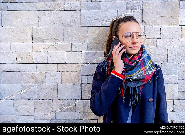 Teenage girl wearing warm clothing talking over smart phone against brick wall at home