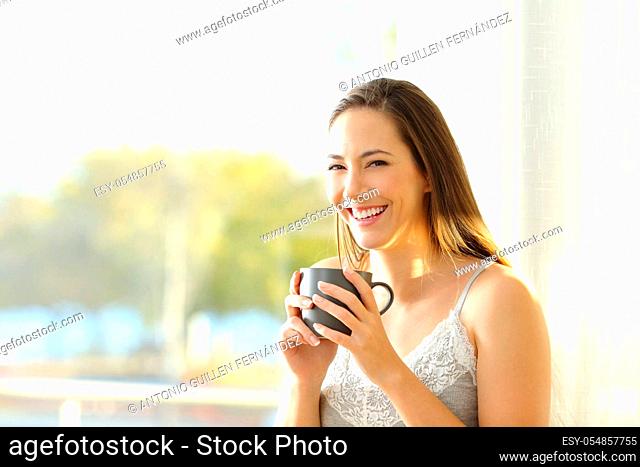 Happy woman at home holding a coffee cup looks at you beside a window in a sunny day