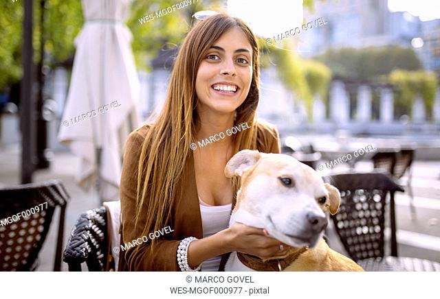 Spain, Gijon, Young woman sitting in pavement cafe, tickling her dog