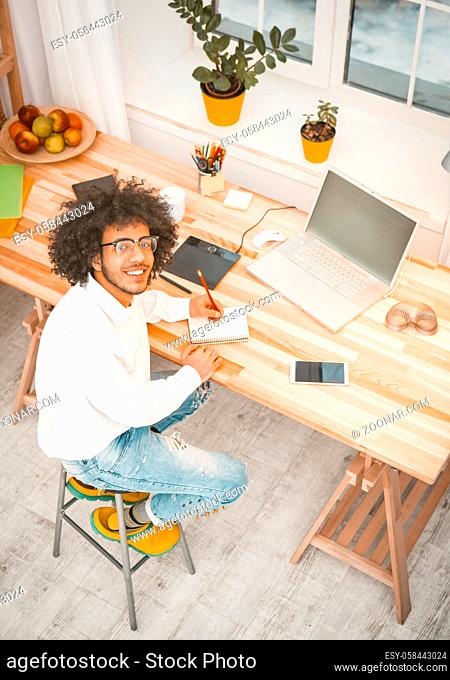 Creative millennial man working at home workplace. Young Arab businessman looks at camera sitting at wooden desk with computer