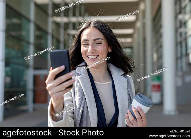 Smiling businesswoman with disposable cup taking selfie through smart phone