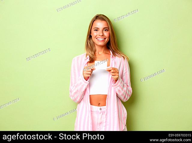 Young beautiful woman with blond hair, showing credit card and smiling happy, standing over green background