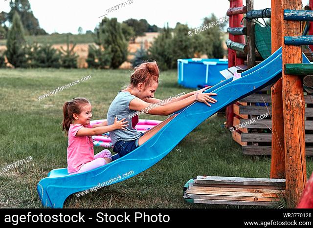 Teenage girl playing with her younger sister in a home playground in a backyard. Happy smiling sisters having fun on a slide together on summer day