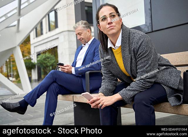 Male and female commuters with smart phones sitting on bench while waiting at bus stop