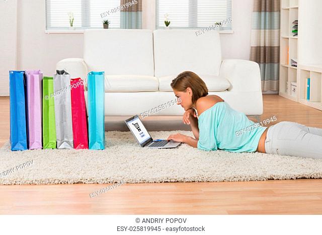 Young Woman Shopping Online On Laptop While Lying On Carpet At Home
