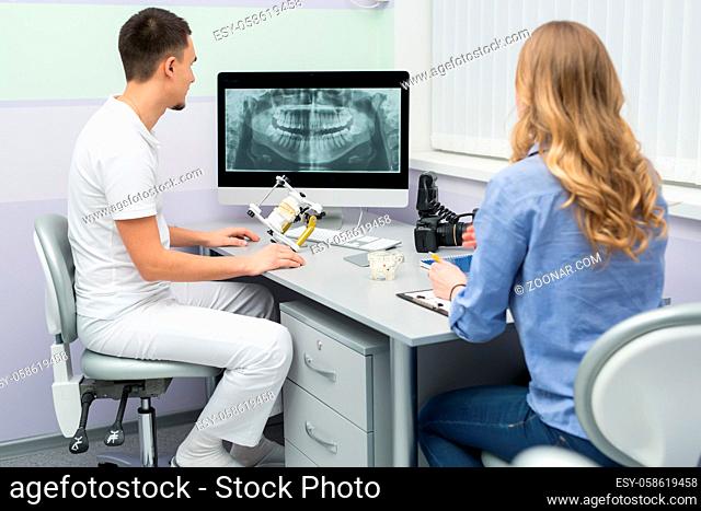 Young dentist in a white uniform and a patient in a blue shirt and jeans sit on chairs at the table. They look at the results of orthopantomogram at the monitor