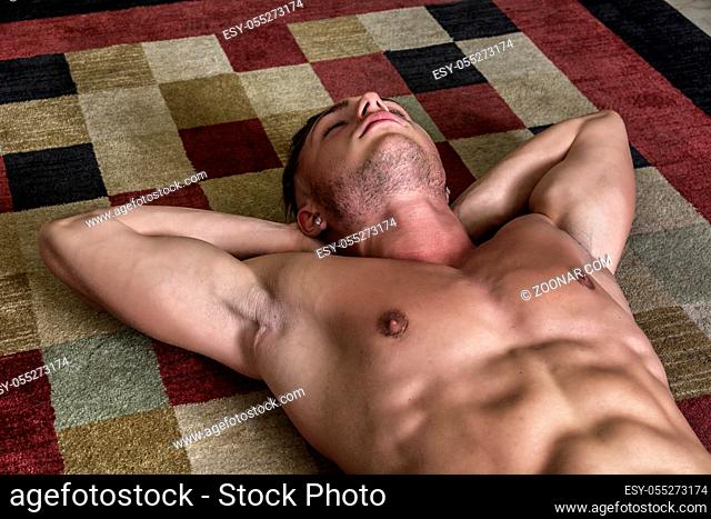 Cool dark-haired shirtless muscular handsome young man laying on the floor over colorful rug, looking at camera, hand under his head