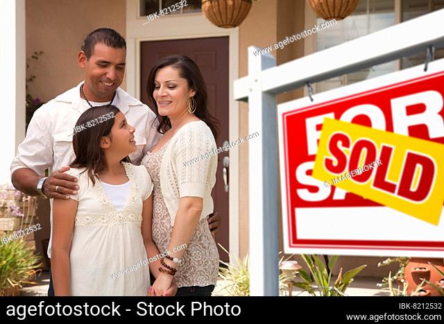 Hispanic mother, father and daughter in front of their new home with sold home for sale real estate sign