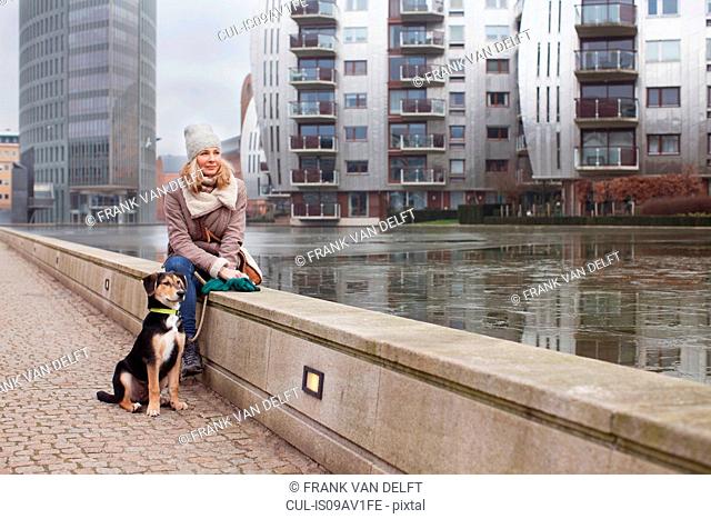 Mid adult woman and her dog sitting on riverside wall