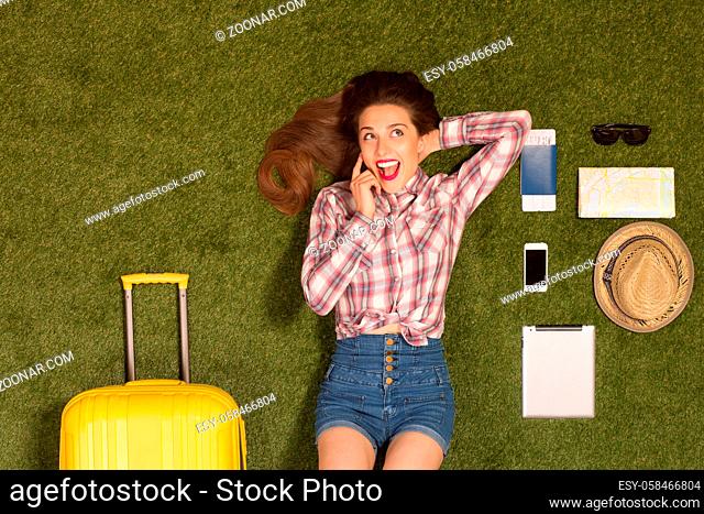 Portrait of happy beautiful tourist lady lying on green grass and talking over mobile or smart phone. The following things are represented near her: map, cap