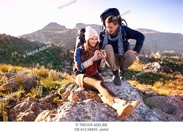 Young couple with backpacks hiking, resting on rock using smart phone