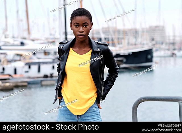 Young woman wearing jacket staring while standing with hands in pockets at harbor
