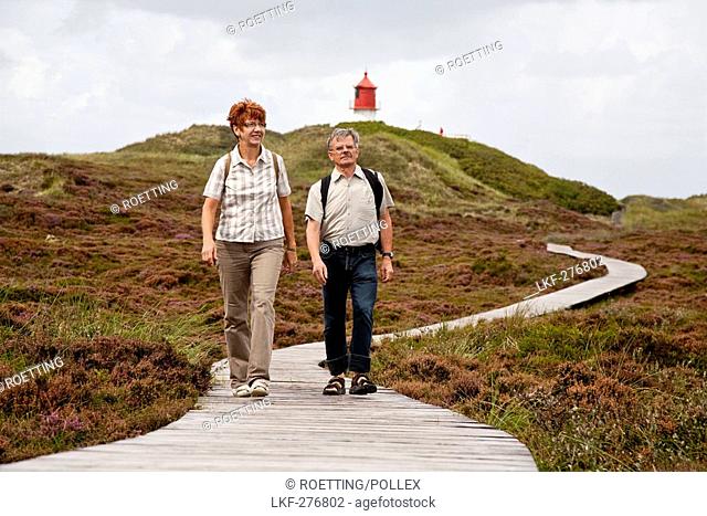 Couple walking along wooden path through dunes, lighthouse in background, Amrum island, Schleswig-Holstein, Germany