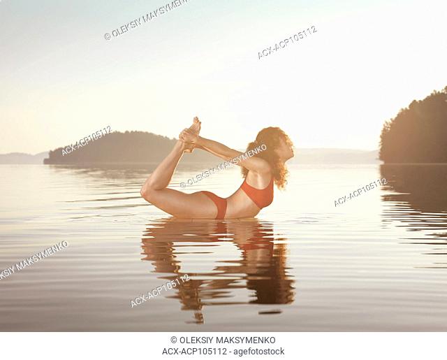 Young woman practicing yoga on a floating platform in water on the lake during misty sunrise in the morning. Yoga Bow posture, Dhanurasana