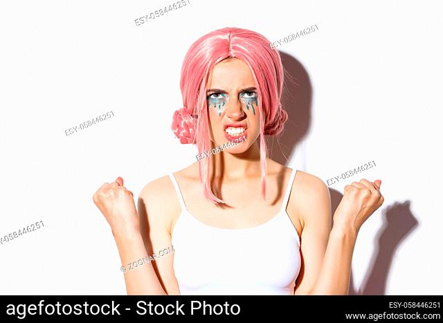 Close-up of angry distressed girl in pink wig, clenching fists and looking up with hatred, standing over white background