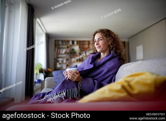 Smiling woman wearing shawl drinking coffee while sitting on sofa at home