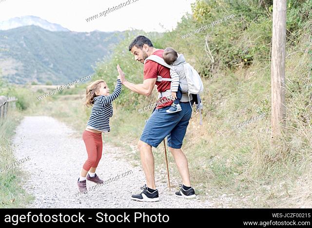 Father hiking with his children, carrying son on back while giving high five to his daughter