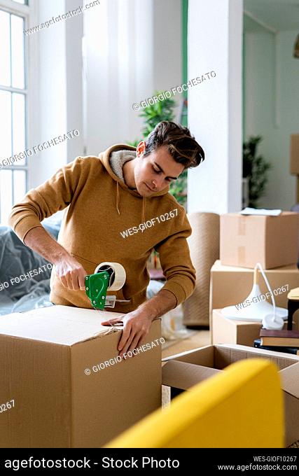 Young man packing cardboard box with adhesive tape in new apartment