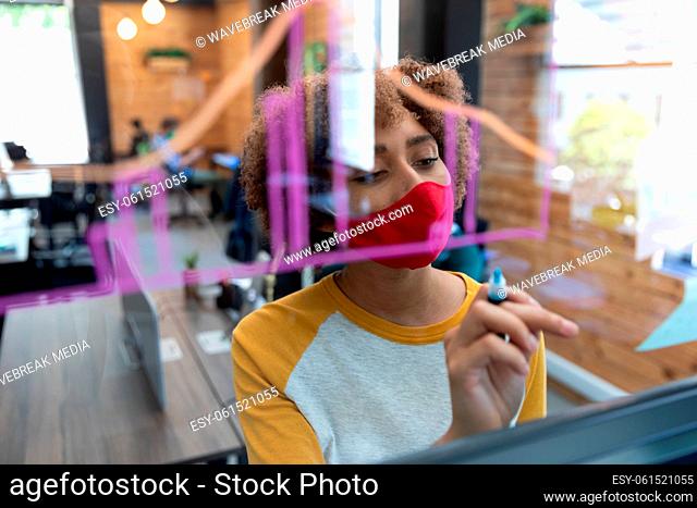 Mixed race woman wearing face mask drawing chart on glass wall in office