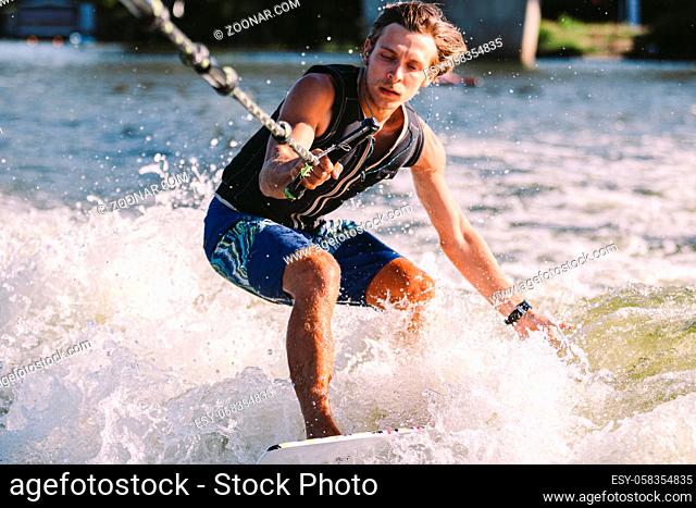 Beautiful long haired blond wakesurf in vest on board along waves of lake. Athletic male athlete wakes surfing in summer on river