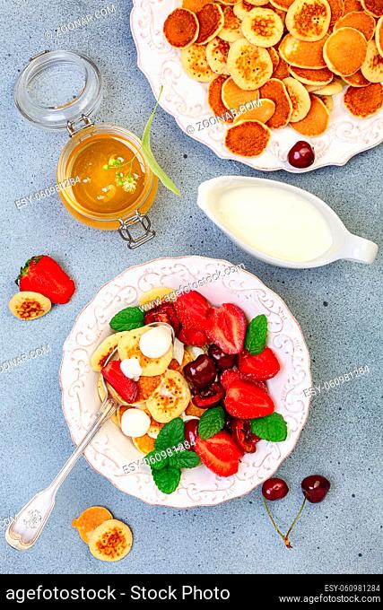 Tiny pancakes with strawberries, cherries, cream and honey in a white bowl and a dish with small pancakes on a textured background. Top view, flat lay