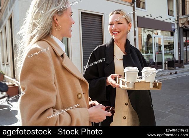 Waist-up portrait of a cheerful mature lady with takeaway coffee conversing with a blonde female