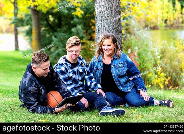 A young man with Down Syndrome sitting with his family and playing games on a tablet, while enjoying each other's company in a city park on a warm fall evening;...