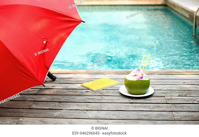 Coconut juice with flower and book and red umbrella by the swimming pool