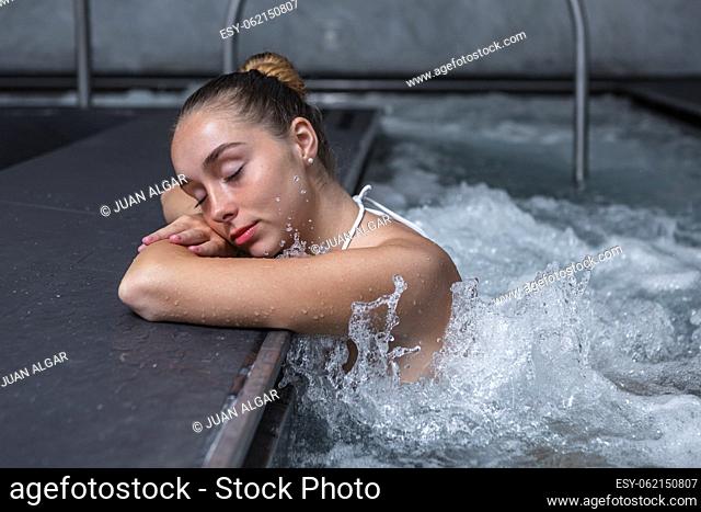 Relaxed young woman closing eyes and holding head on crossed arms while chilling in splashing water of massage pool on weekend day on spa resort