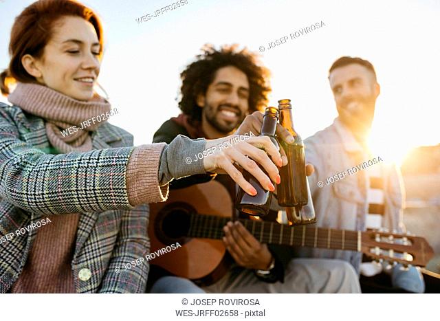 Three happy friends with guitar toasting beer bottles at sunset