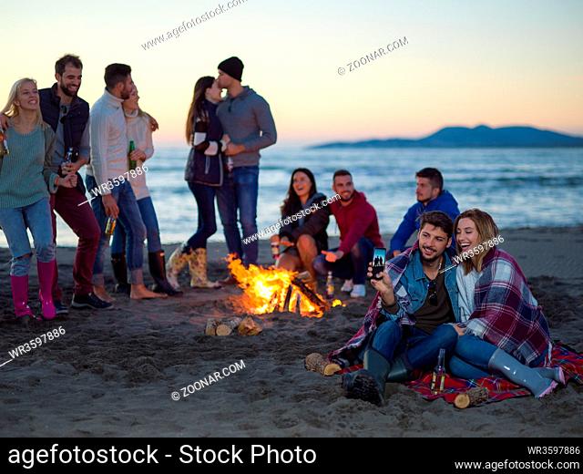 Couple using cell phone during autumn beach party with friends drinking beer and having fun