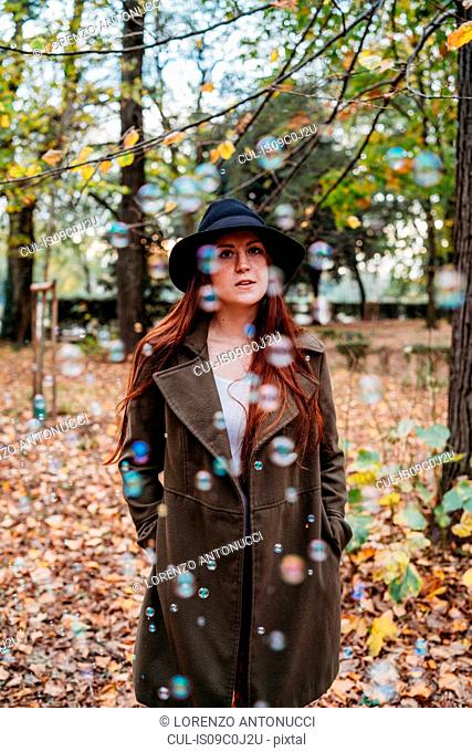 Young woman with long red hair amongst floating bubbles in autumn park, portrait