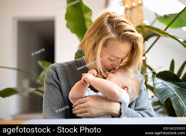 Portrait of young mother cuddling and kissing her adorable little baby boy while sitting at the table at home. Sensory stimulation for baby development