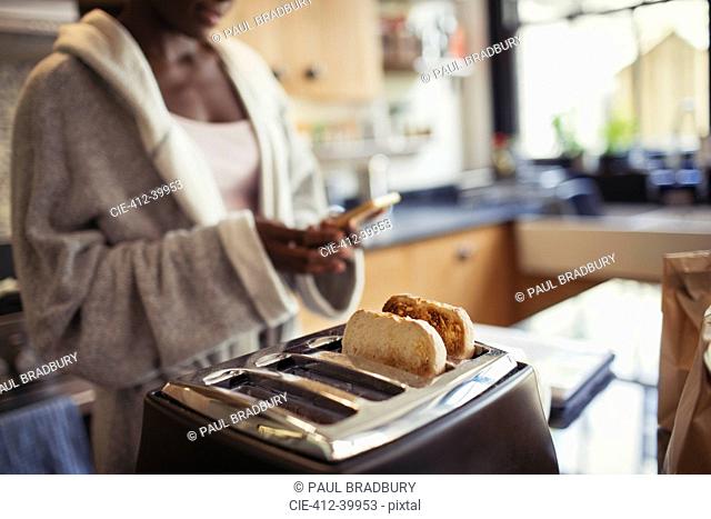 Woman texting with smart phone, toasting bread in toaster in kitchen