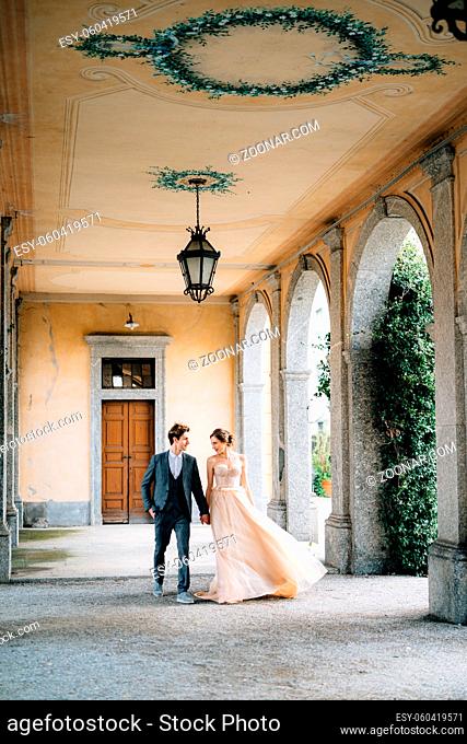 Bride and groom walk along the old terrace with columns entwined with green ivy. Lake Como, Italy. Front view. High quality photo