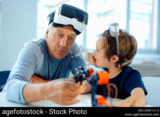 Senior man with VR glasses examining robot model by grandson at home