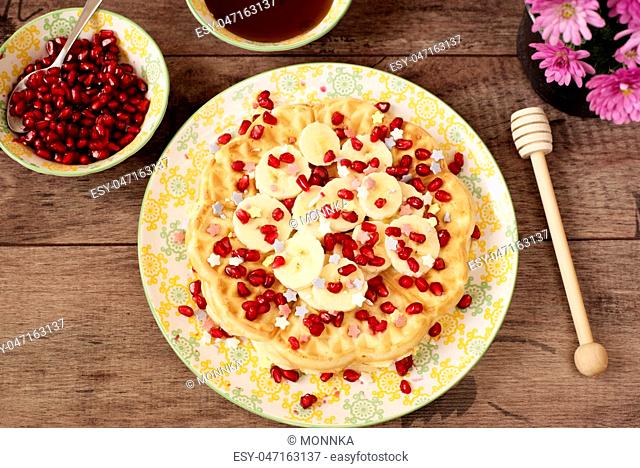 Soft Belgian heart shaped waffles with banana and pomegranate, sugar decoration stars, covered with honey. Rustic wooden background, top view, close-up