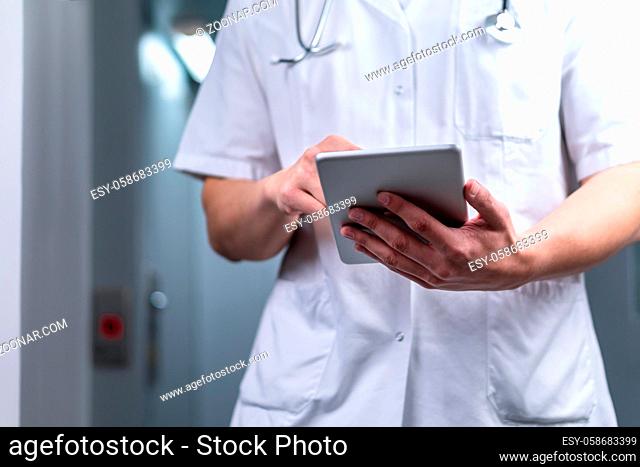 photo of male doctor in uniform with stethoscope coming out of the elevator and using computer tablet in hospital. Modern medical concept