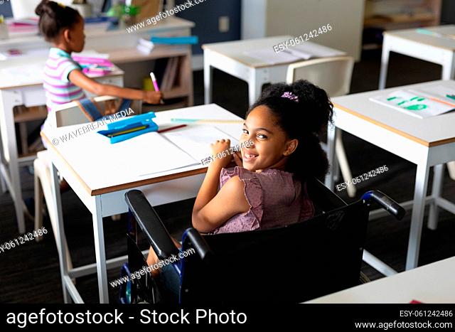 Portrait of happy biracial elementary schoolgirl sitting on wheelchair at desk in classroom