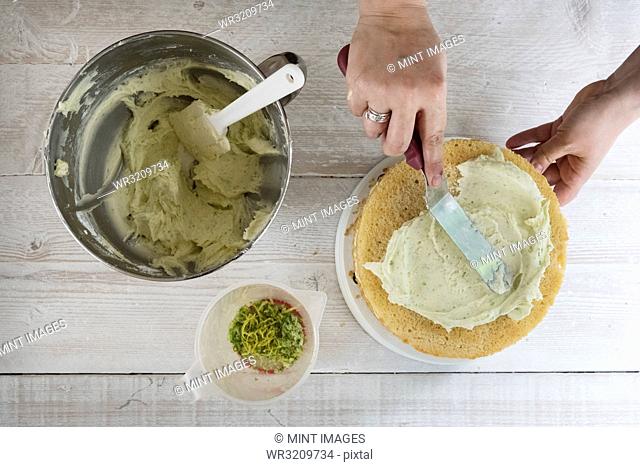 A fresh gin and tonic flavoured baked cake being iced, a hand with a palette knife smoothing icing, and a bowl of fresh icing and lemon zest